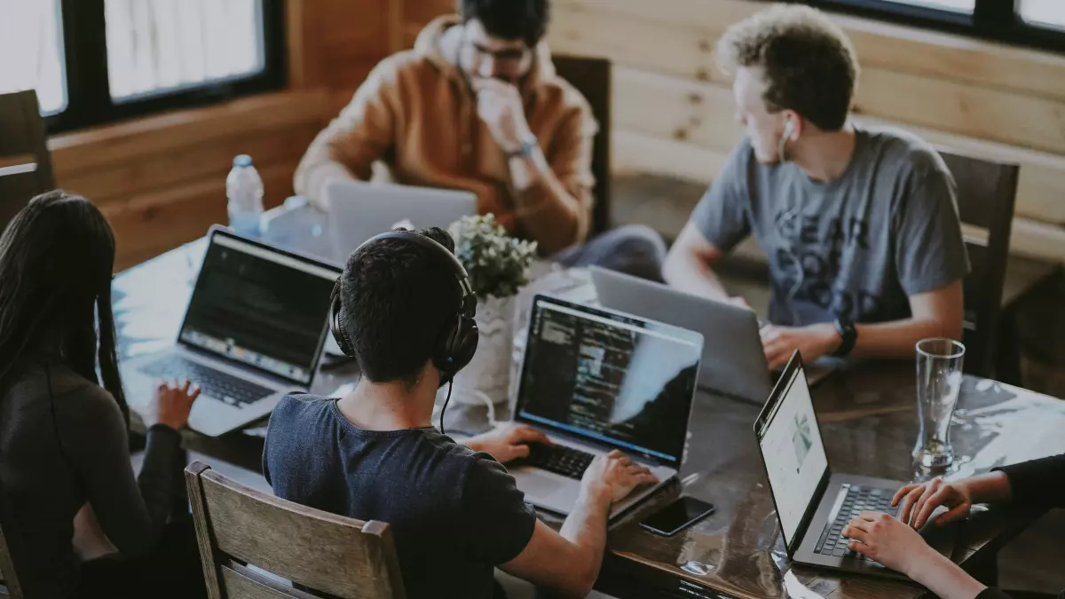 A team of four young professionals are working together at a table, each with a laptop open in front of them.