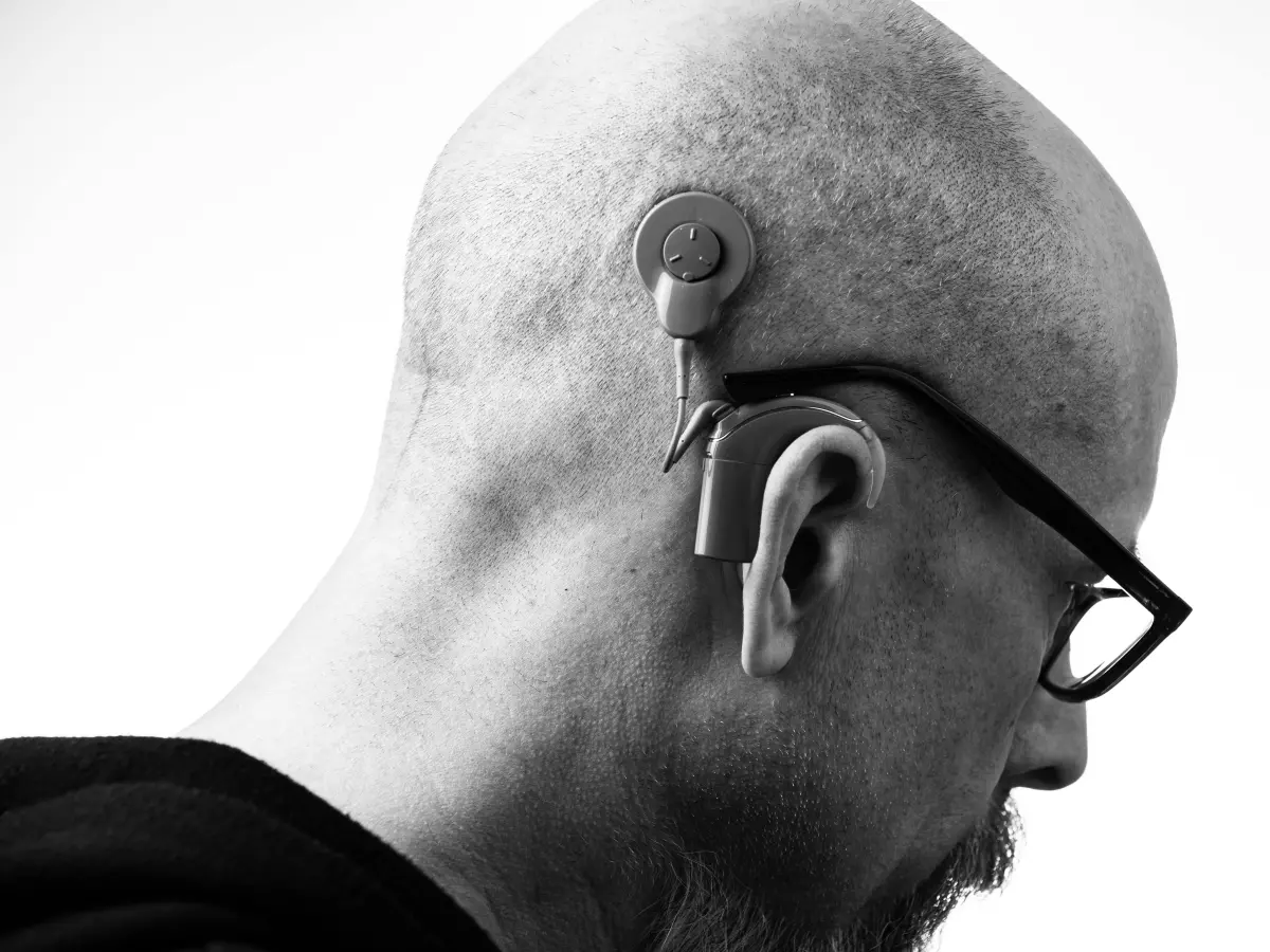 A man is wearing a hearing aid in his right ear. He is looking down and his hair is bald.