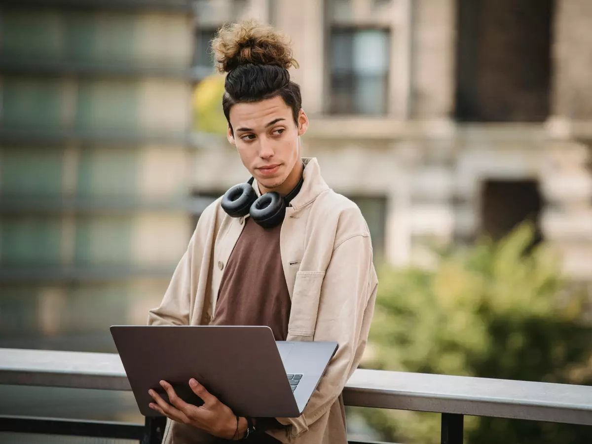 A young person in a beige shirt and grey sweatshirt, is standing on a balcony, looking at a laptop, they have their headphones around their neck