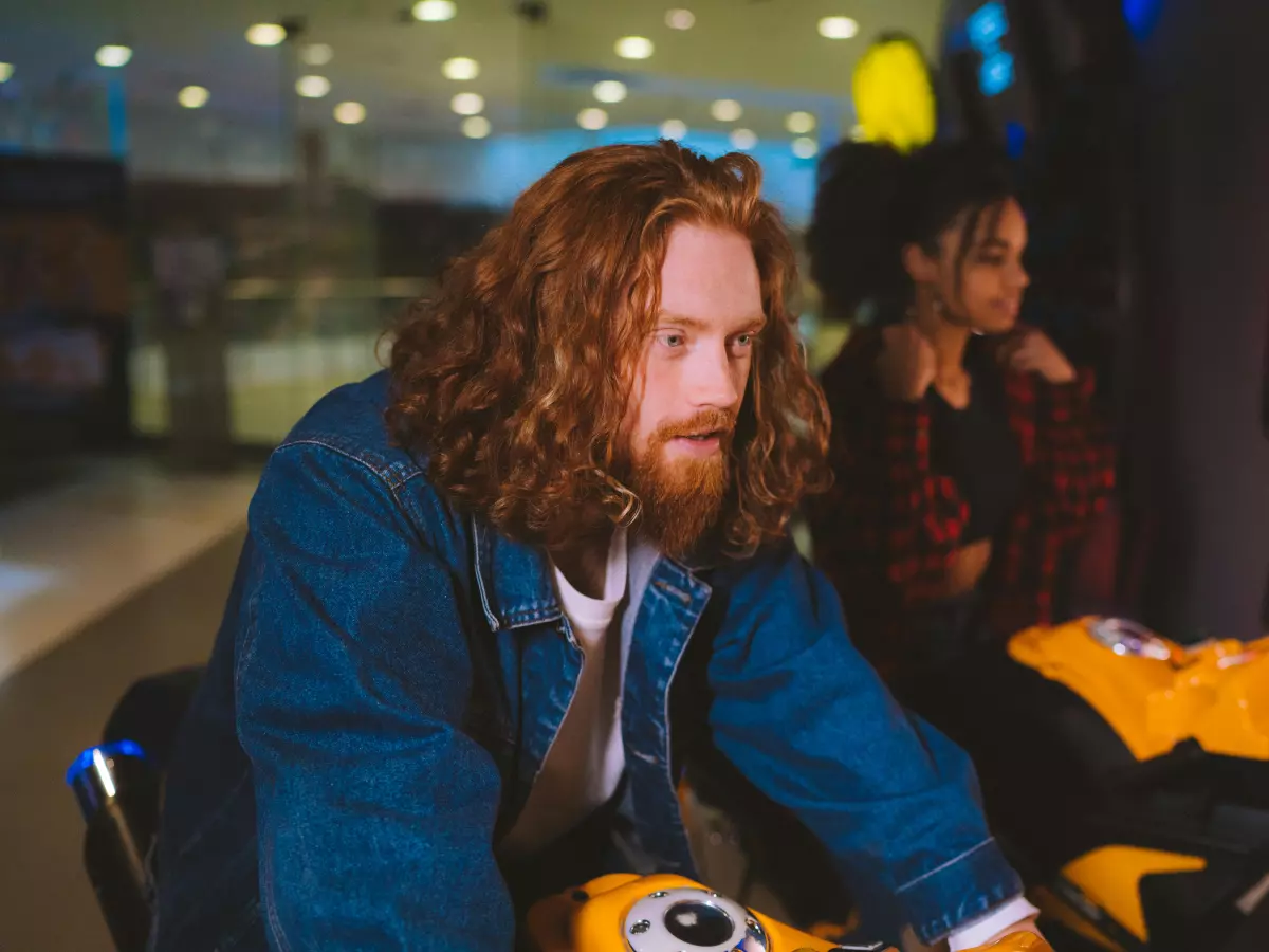 A young man with long red hair is sitting at an arcade machine, wearing a blue denim jacket and a white T-shirt. His focus is on the game as he enjoys playing. 