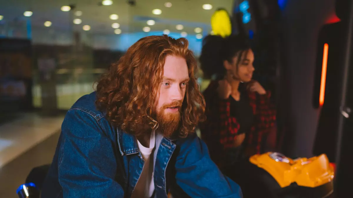 A young man with long red hair is sitting at an arcade machine, wearing a blue denim jacket and a white T-shirt. His focus is on the game as he enjoys playing. 