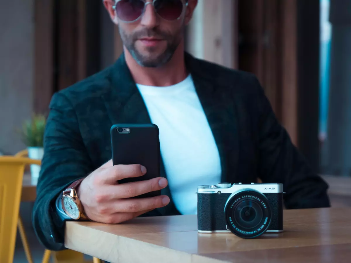 A man is sitting at a table with a smartphone in one hand and a professional camera on the table in front of him.