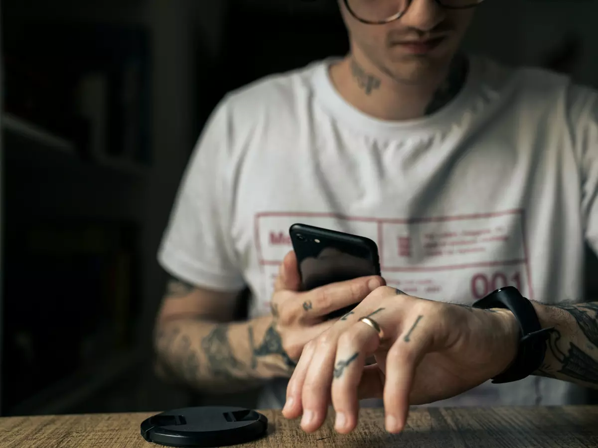 A close-up shot of a man wearing a smartwatch and holding a smartphone. He has tattoos on his arms and is wearing a white t-shirt.