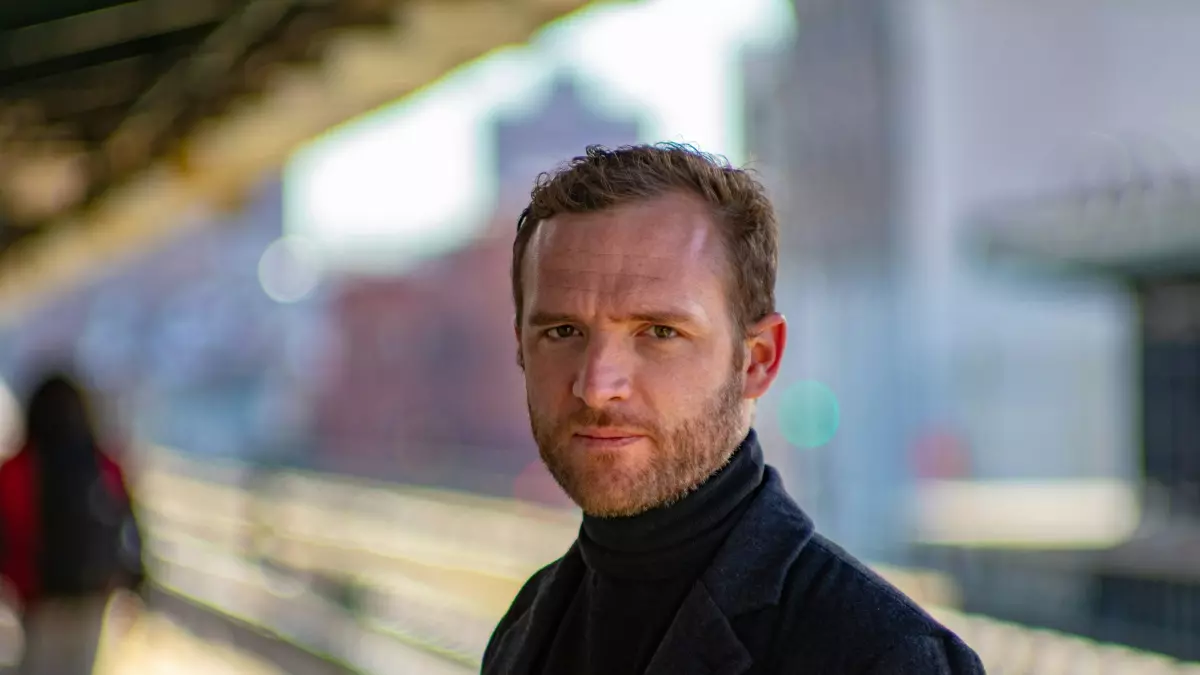 A man standing on a train platform, looking directly at the camera, wearing a black coat and a turtleneck sweater, with a blurry background of a city street.