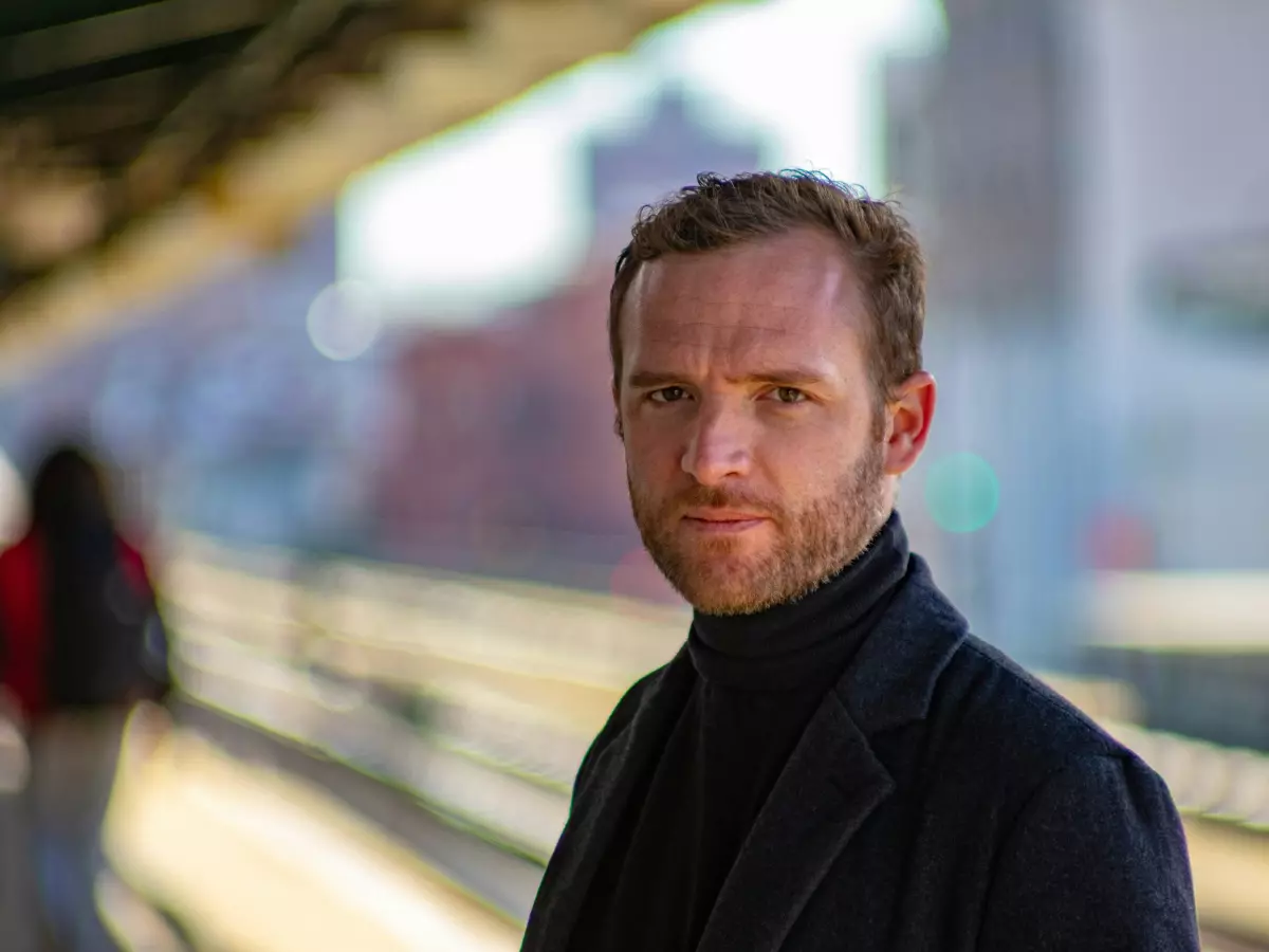 A man standing on a train platform, looking directly at the camera, wearing a black coat and a turtleneck sweater, with a blurry background of a city street.