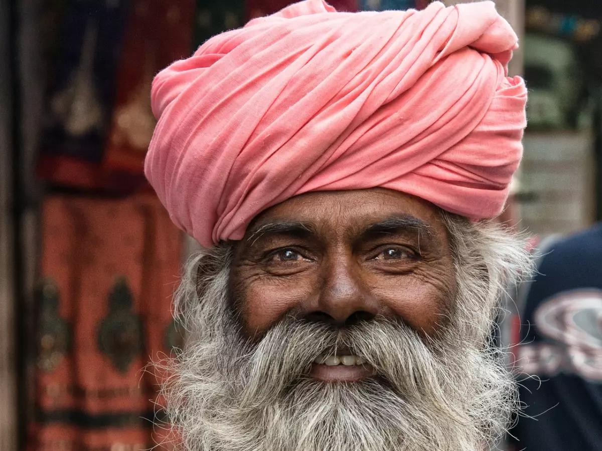 A portrait of a man with a long white beard and mustache, wearing a pink turban. He is smiling and looking at the camera.