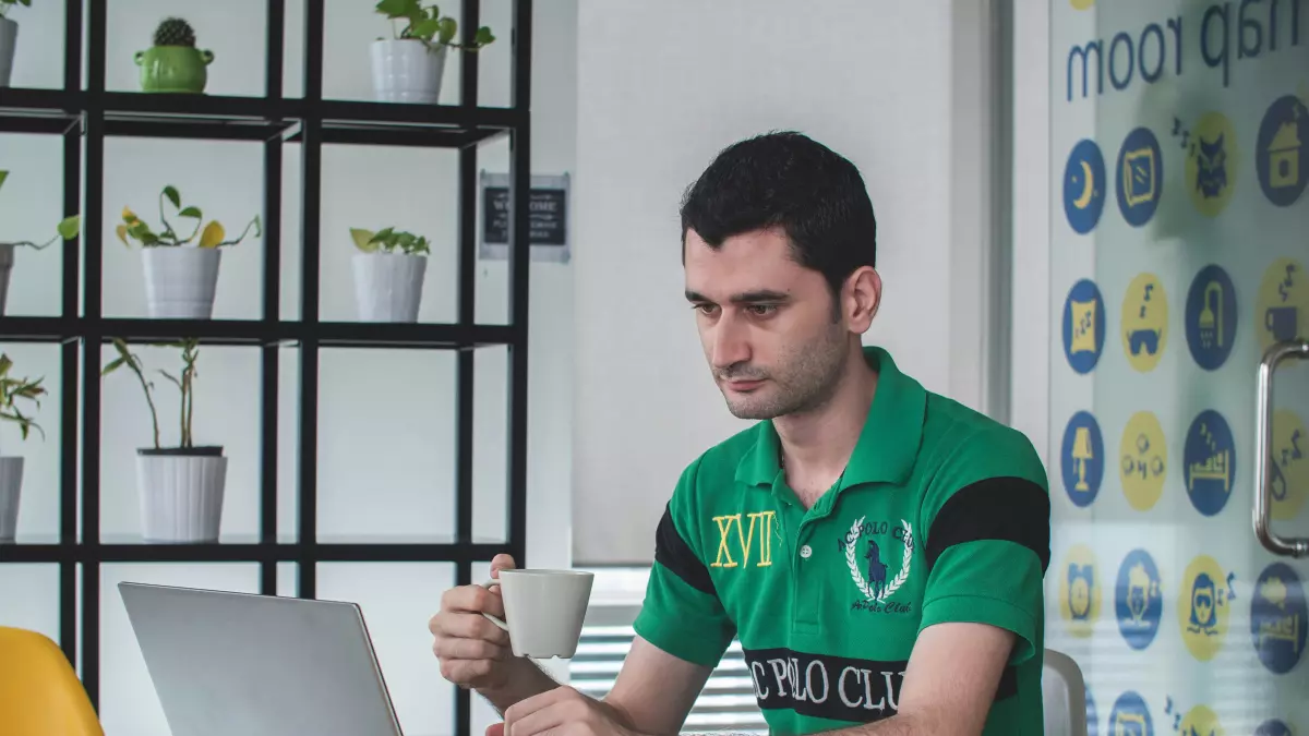 A man sitting at a table working on his laptop, with a cup of coffee in his hand. He is dressed casually and appears focused on his work.