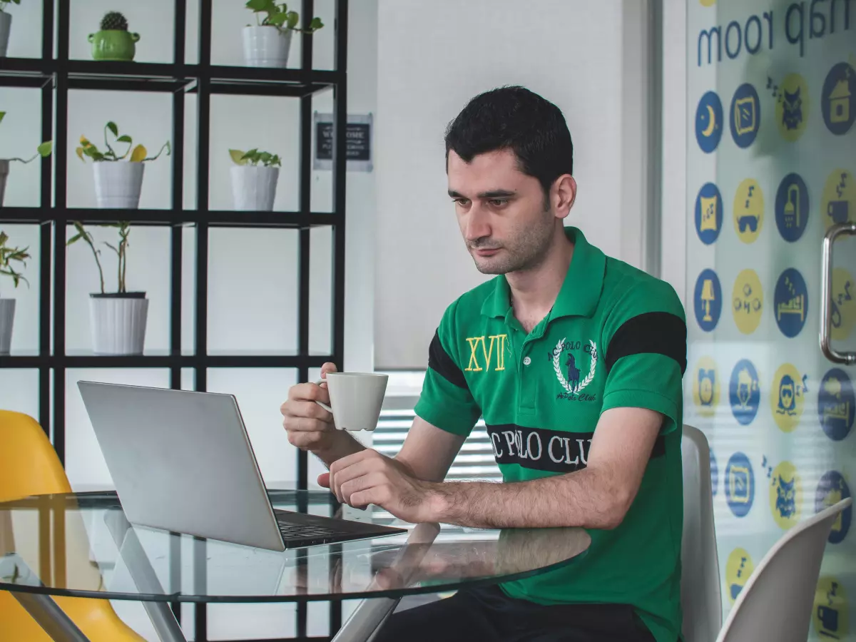 A man sitting at a table working on his laptop, with a cup of coffee in his hand. He is dressed casually and appears focused on his work.