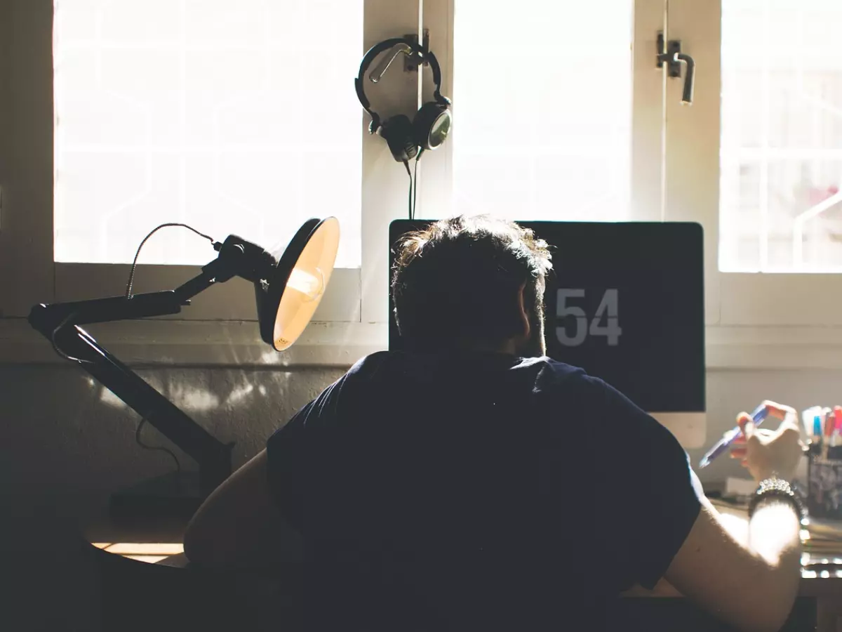 A person sitting at a desk with a computer, looking at the screen. The person is wearing headphones and there is a lamp on the desk.