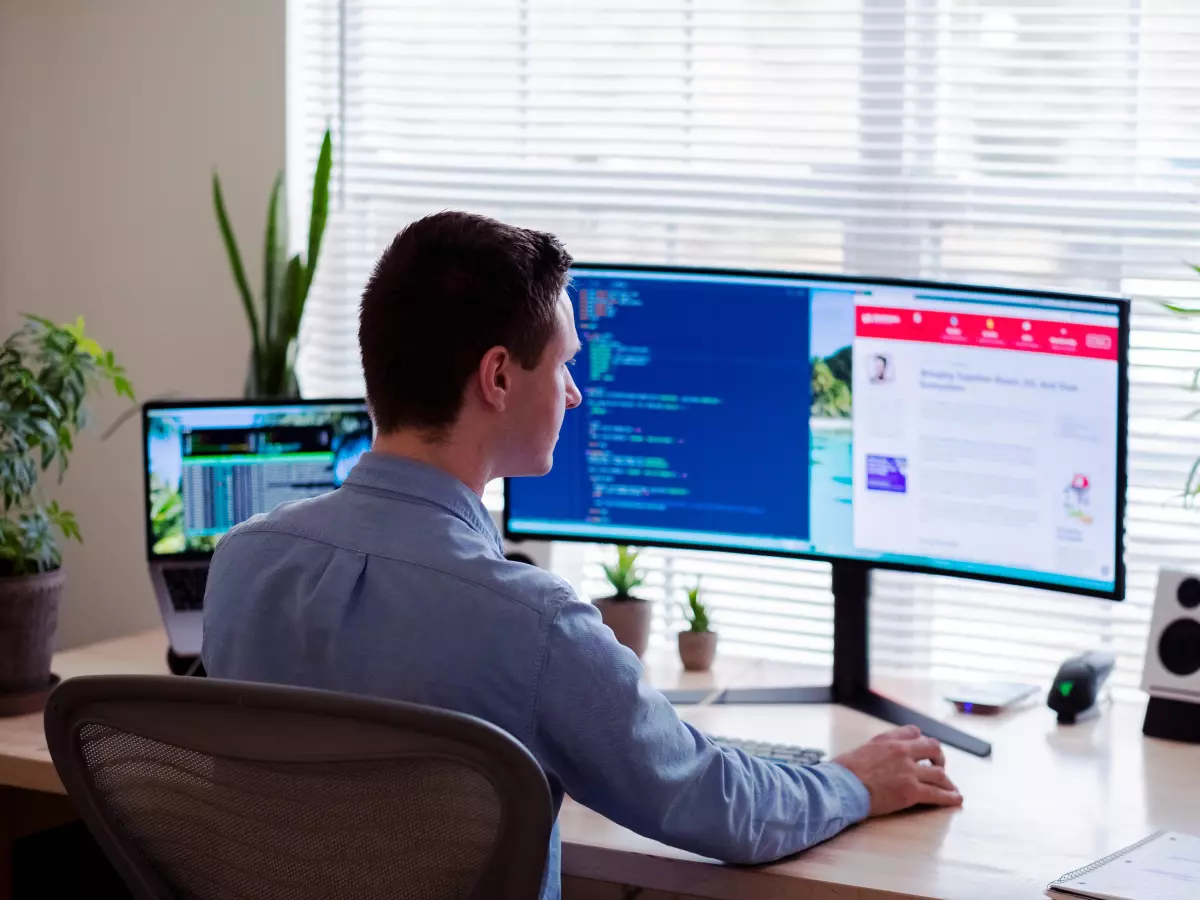 A man in a blue shirt sits at a desk with two large computer monitors in front of him. He's using a keyboard and mouse. The background shows a window, plants, and another laptop, emphasizing a home office setting. 