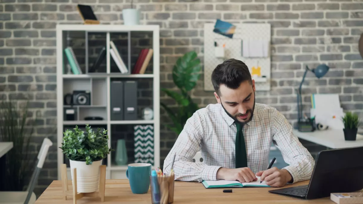 A man sitting at a desk in an office, working on a laptop and taking notes.