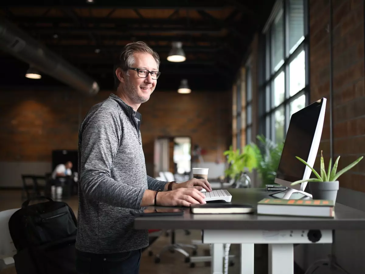 A man in a grey sweater and glasses is standing at a desk working on a computer in a bright office.