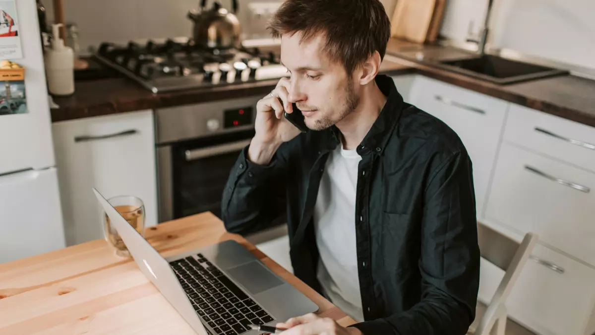 A man in a black shirt sits at a table in a kitchen and talks on his phone while looking at a laptop. There is a cup of coffee in front of him. The background is a kitchen with a white stove, oven and cabinets.