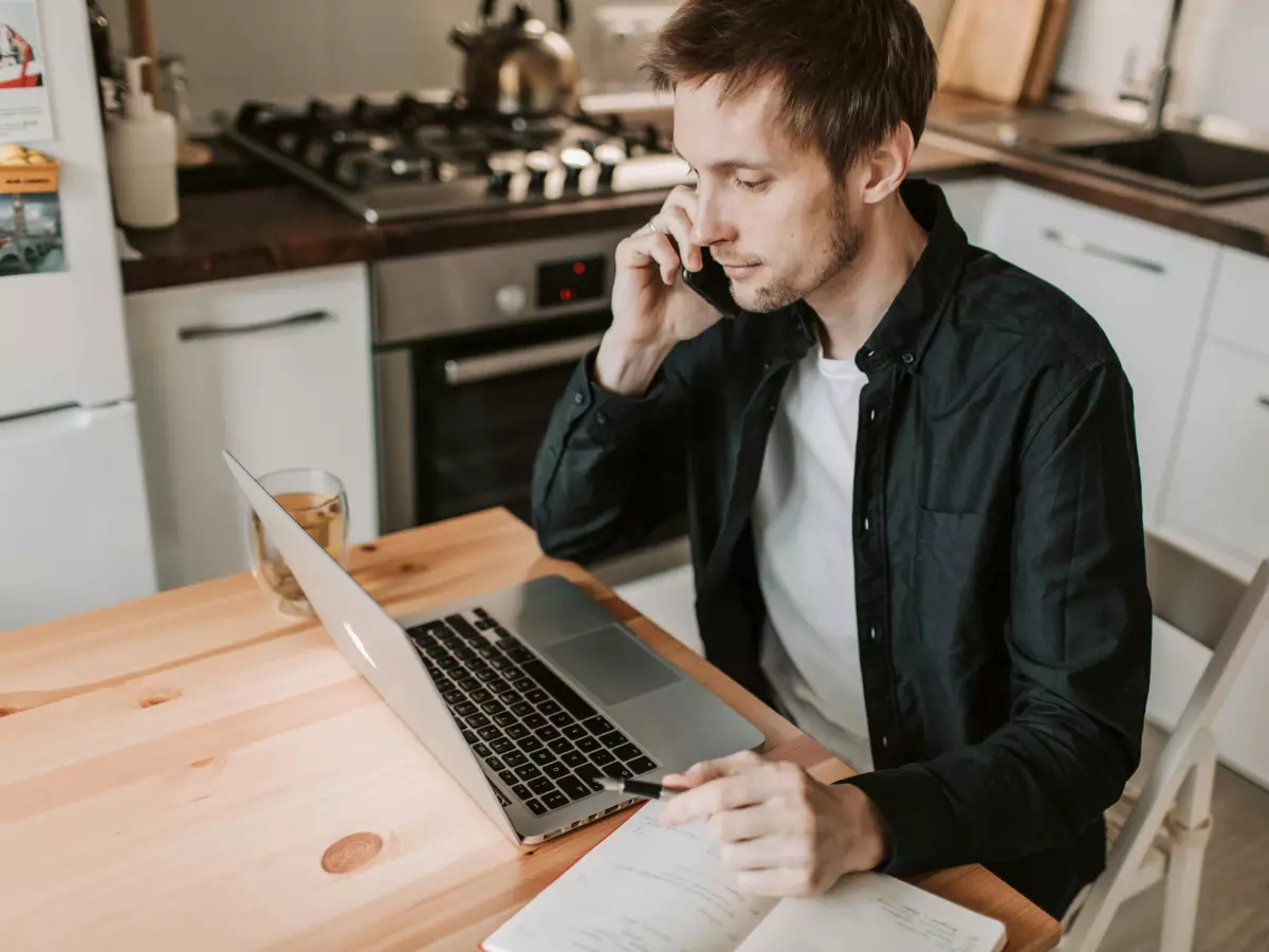 A man in a black shirt sits at a table in a kitchen and talks on his phone while looking at a laptop. There is a cup of coffee in front of him. The background is a kitchen with a white stove, oven and cabinets.