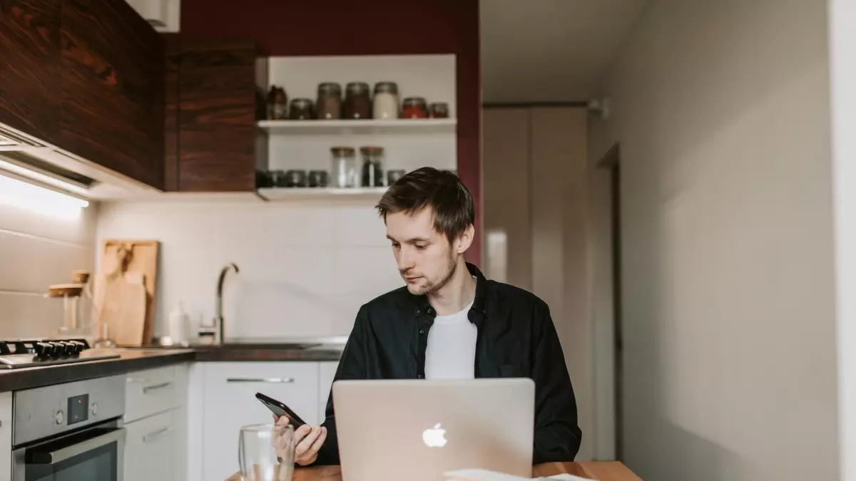 A man sits at a table in his kitchen, using a laptop and holding a phone. The kitchen is modern and has a minimalist design.