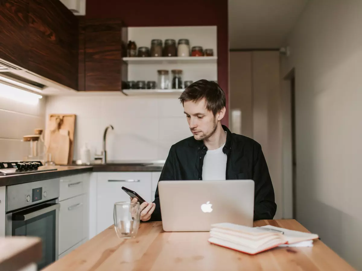 A man sits at a table in his kitchen, using a laptop and holding a phone. The kitchen is modern and has a minimalist design.