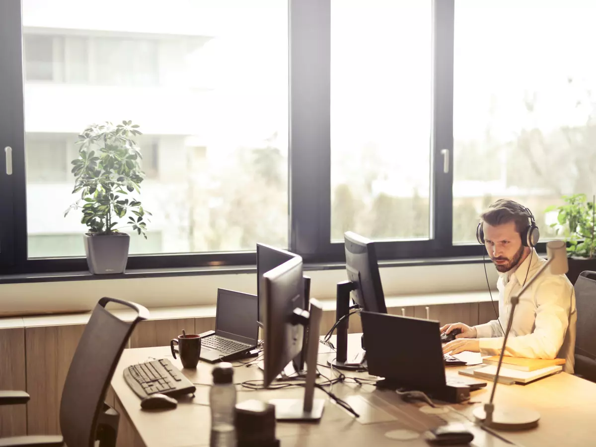 A man in a white shirt and headphones is working on a laptop computer in an office setting. He is sitting at a desk with several other computers behind him. There is a plant and a water bottle on the desk.