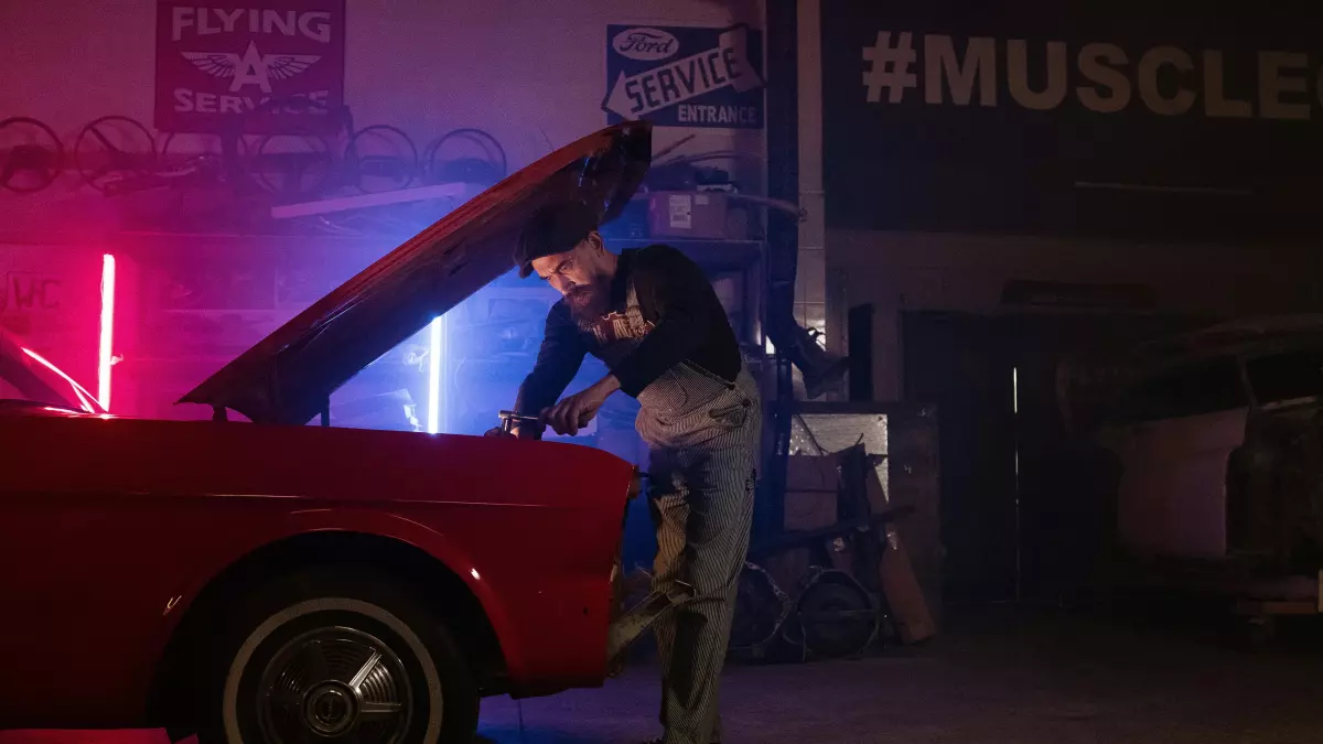 A man is working on a red classic car in a garage, the hood is open, and neon lights create a dramatic atmosphere.