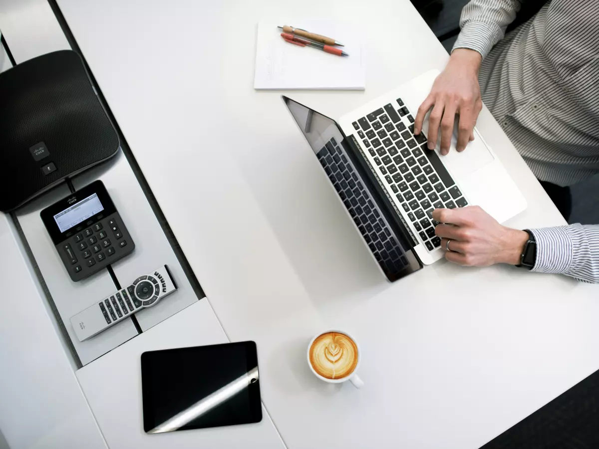 A person is working on a laptop at a white desk. The laptop is open and the person's hands are typing on the keyboard. There is a cup of coffee on the desk.