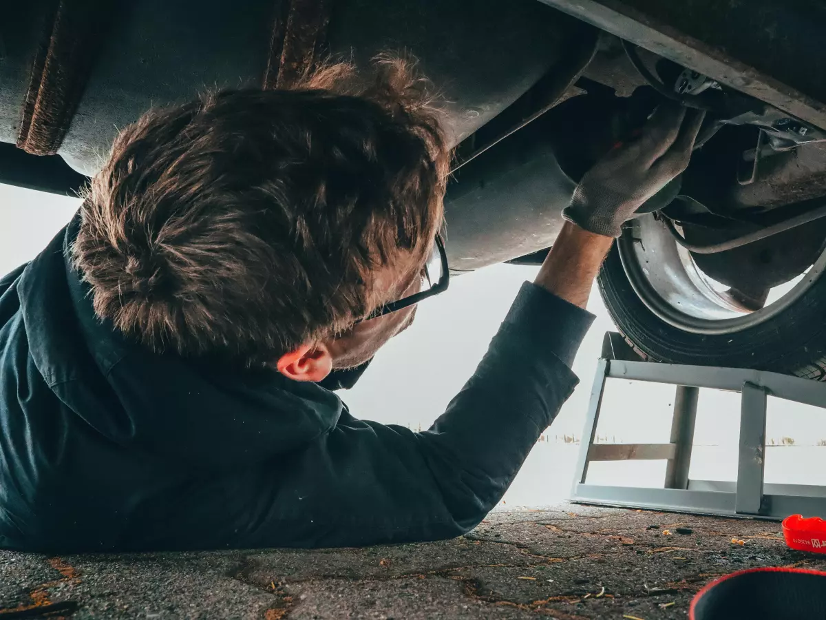 A man in a garage is working on a car. He is wearing a dark shirt and black pants. He is lying on his back under the car with his legs out.