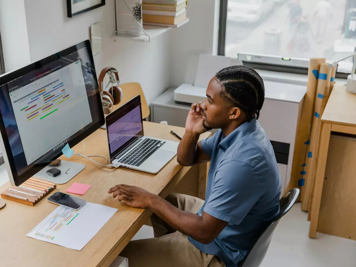 A man sitting at a desk using a laptop and a desktop computer. He is looking at the laptop and talking on the phone.