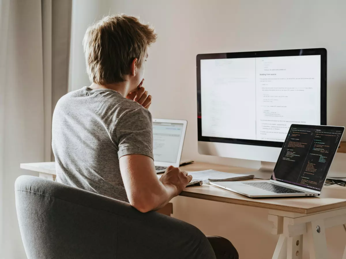 A young man sits at a desk facing two computer screens, one a large monitor displaying code. He is looking intently at the screen with his hand on his chin, appearing focused and contemplative. This image captures the essence of software development and the meticulousness required for effective code composition.