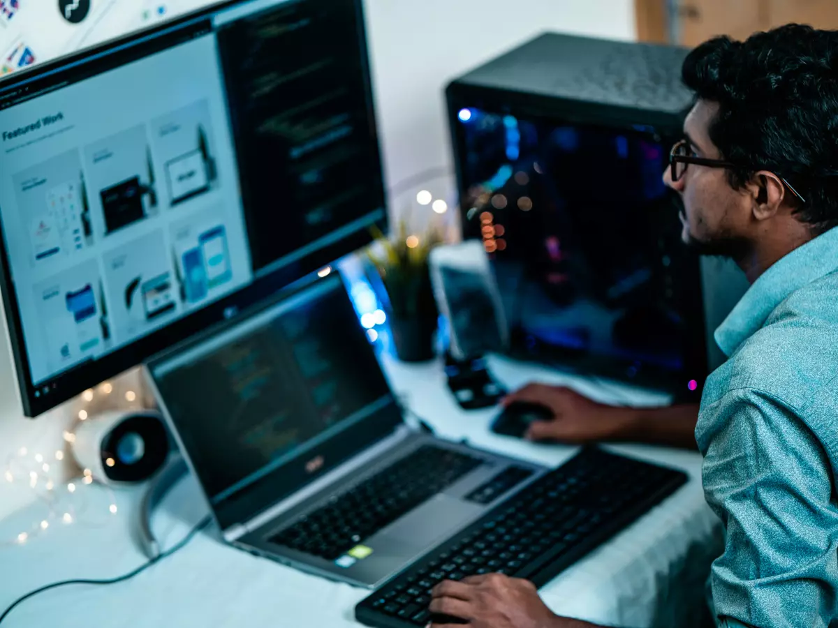 A man is sitting at a desk in front of a computer, laptop and monitor. He is looking at the monitor. The monitor has a website on it, with multiple images in a grid.