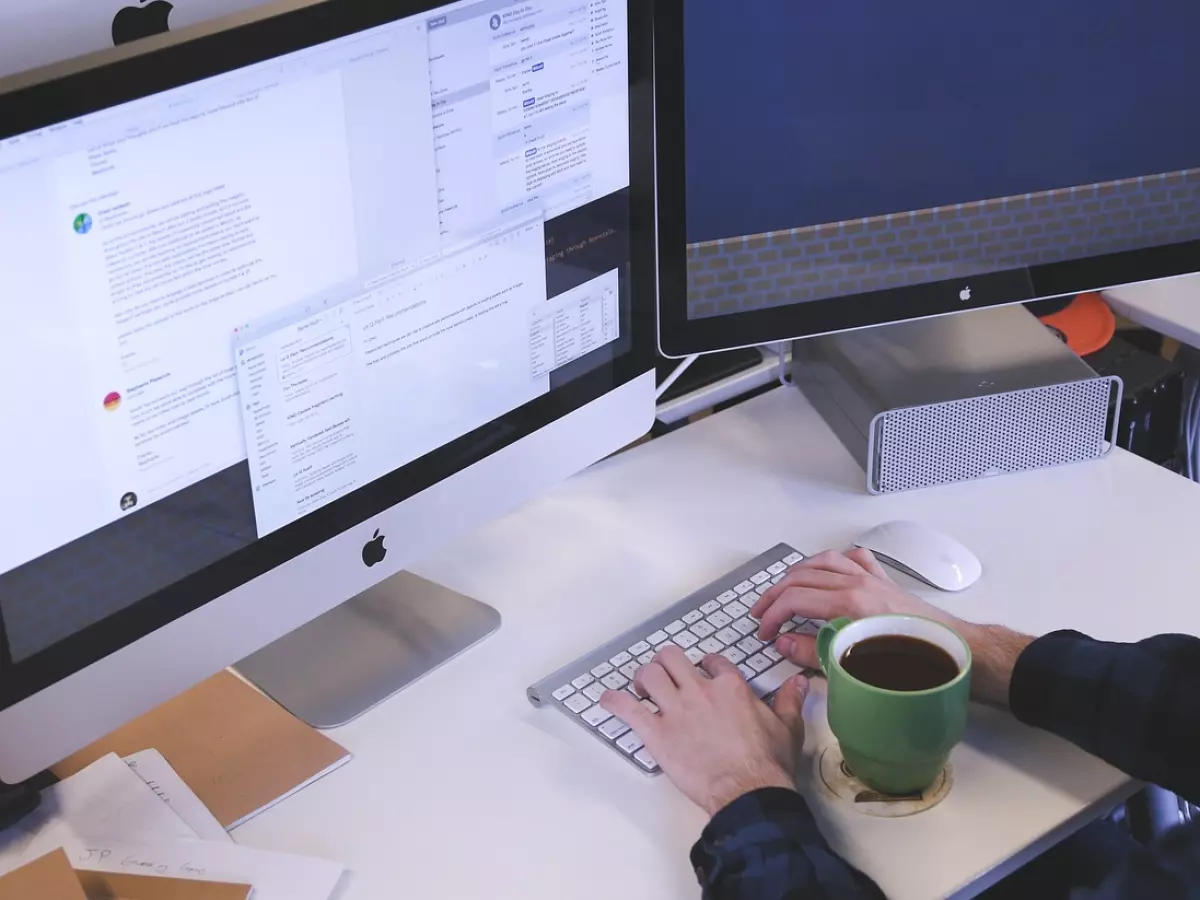 A person is sitting at a desk, using a Mac computer with a green mug of coffee beside it, in front of a large screen.