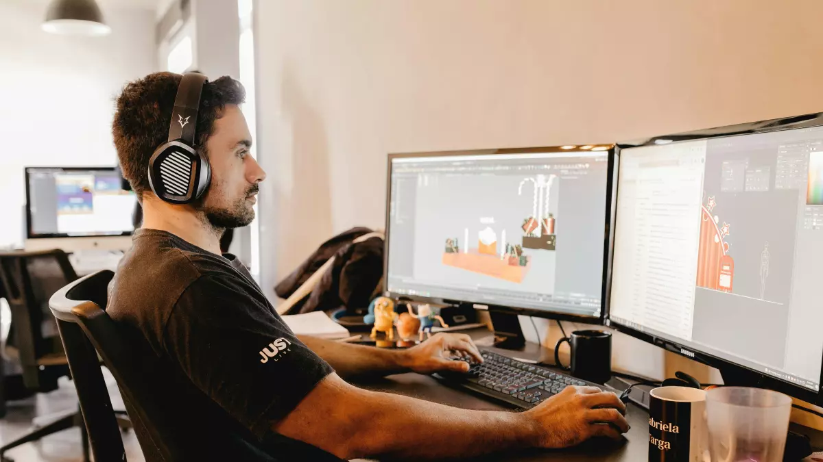 A person working on a computer with dual monitors, wearing headphones and a black t-shirt. The workspace is clean and modern.