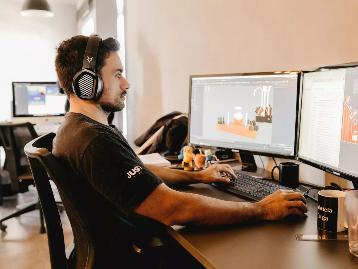 A person working on a computer with dual monitors, wearing headphones and a black t-shirt. The workspace is clean and modern.