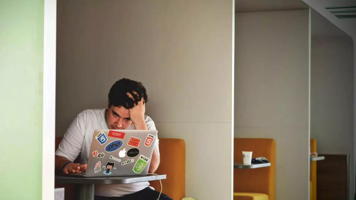 A man sits at a desk in front of a laptop, looking frustrated.