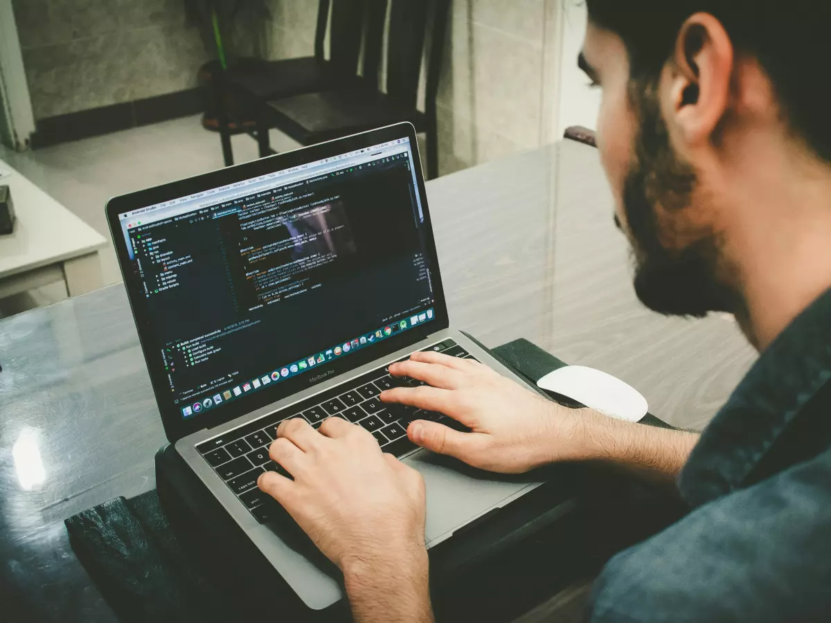 A man sitting at a desk, looking at a laptop screen that displays lines of code. The image is taken from the side, showing his profile and the laptop.