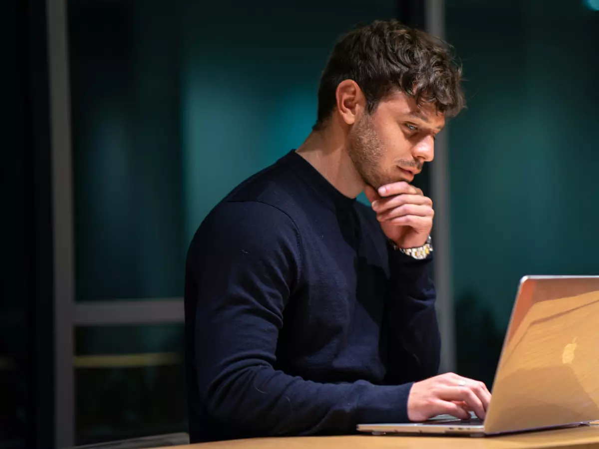 A young man with short brown hair and a beard sits at a desk, looking intently at a laptop screen. He has his hand on his chin and appears to be thinking deeply. The background is blurred, suggesting an office or work environment.