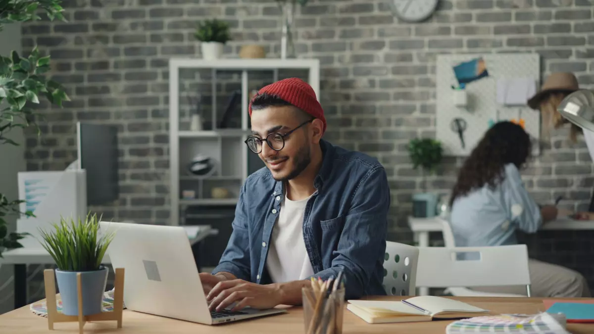 A man sitting at a desk in an office, looking at a laptop screen, wearing a red beanie and denim jacket. The laptop is in the foreground, and the man