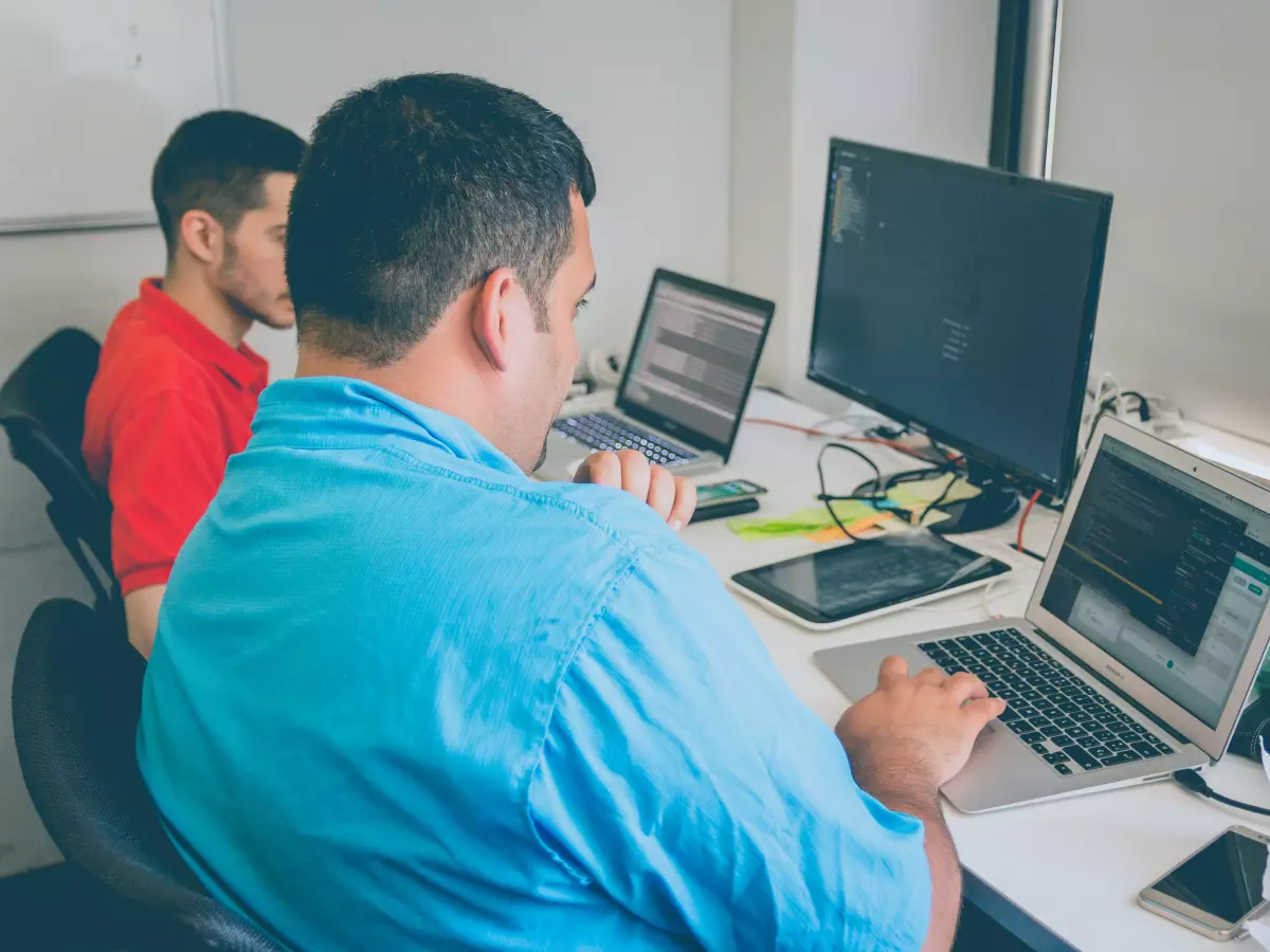 Two men are working on computers in an office. They are looking at their screens, which are filled with code.