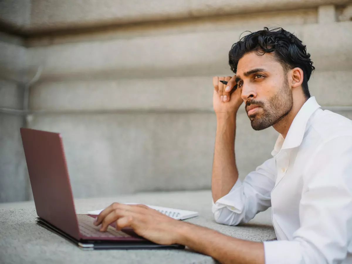 A young man with a beard and a white shirt is sitting on a concrete step, looking frustrated while working on a laptop. He is holding his hand to his forehead, looking tired and stressed. 