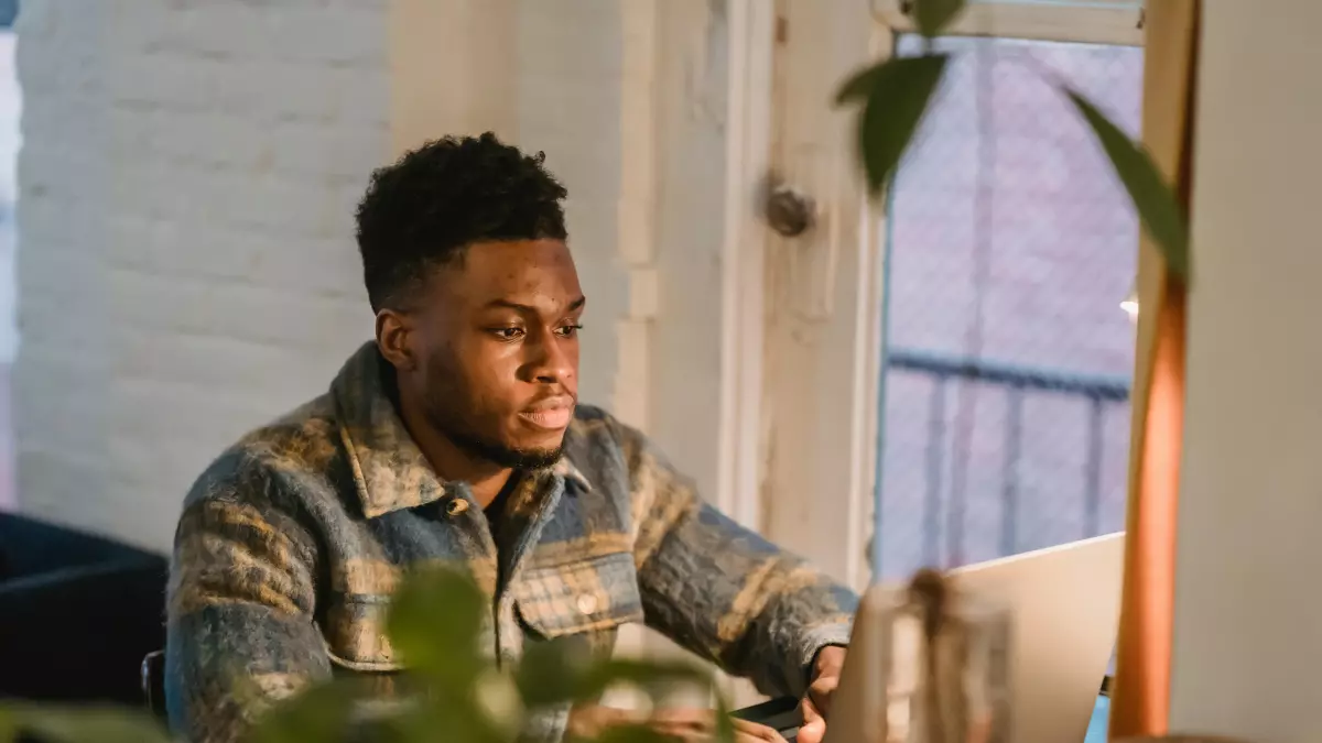 A man sitting at a table in a living room, using a laptop. There are plants and other home decor in the background.