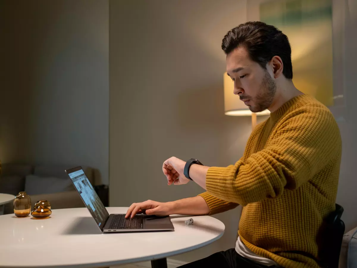 A man wearing a yellow sweater is sitting at a table with a laptop and is checking the time on his Pixel Watch.