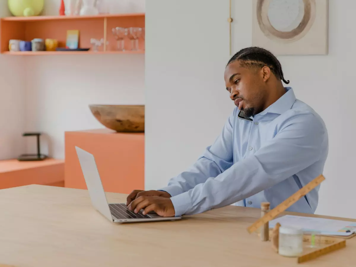 A man is sitting at a table working on his laptop. He is wearing a blue shirt and has his head down, focused on his work.