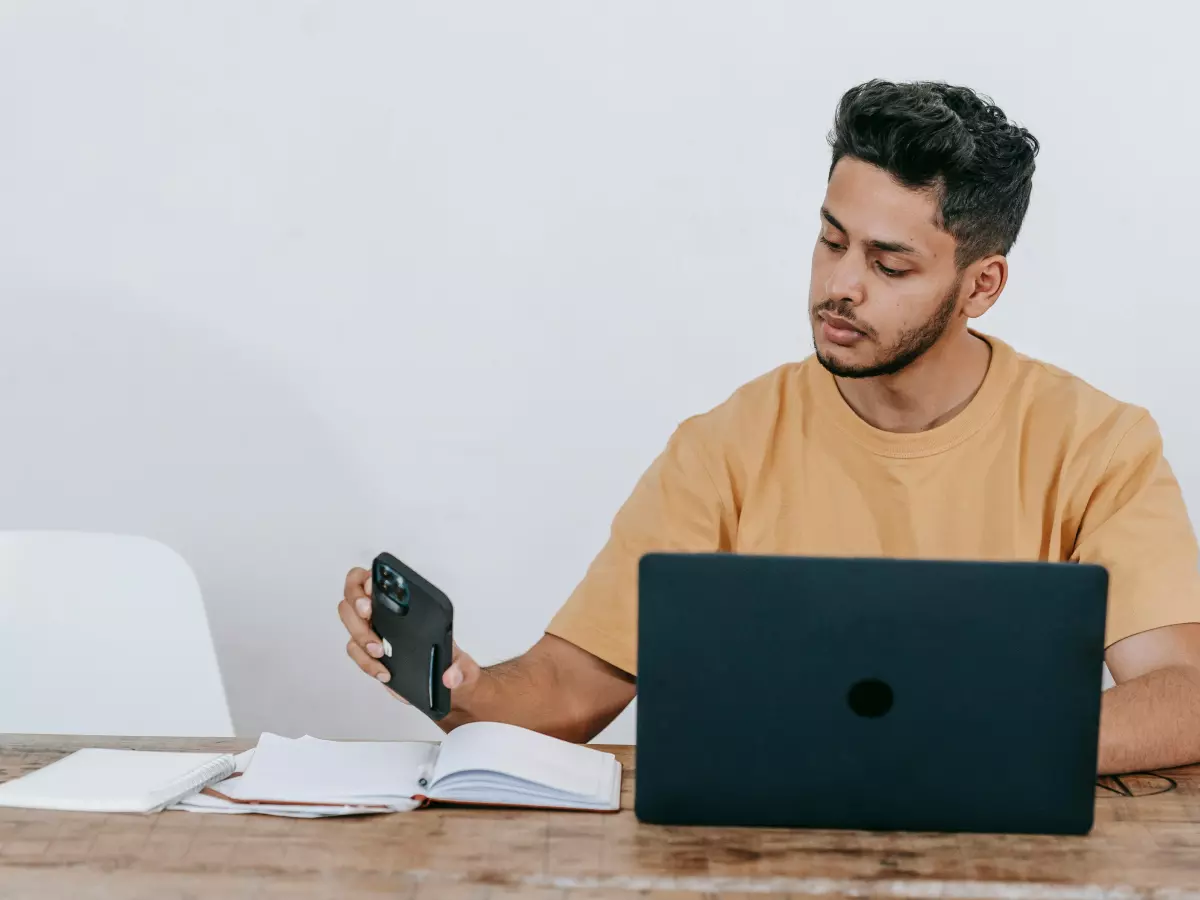 A young man is working on a laptop while looking at his phone.