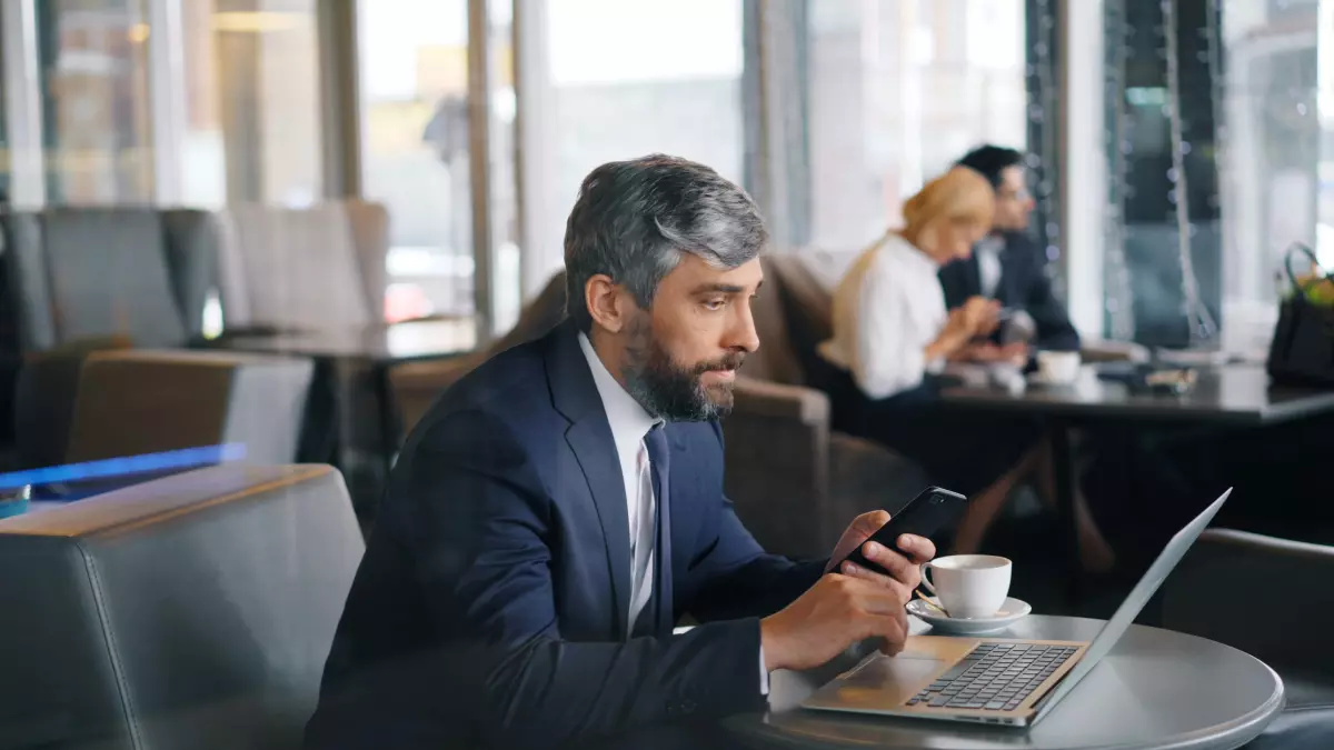 A man in a suit sits at a table in a cafe, looking intently at his phone while a laptop is open in front of him. He is surrounded by other people, suggesting a busy environment.