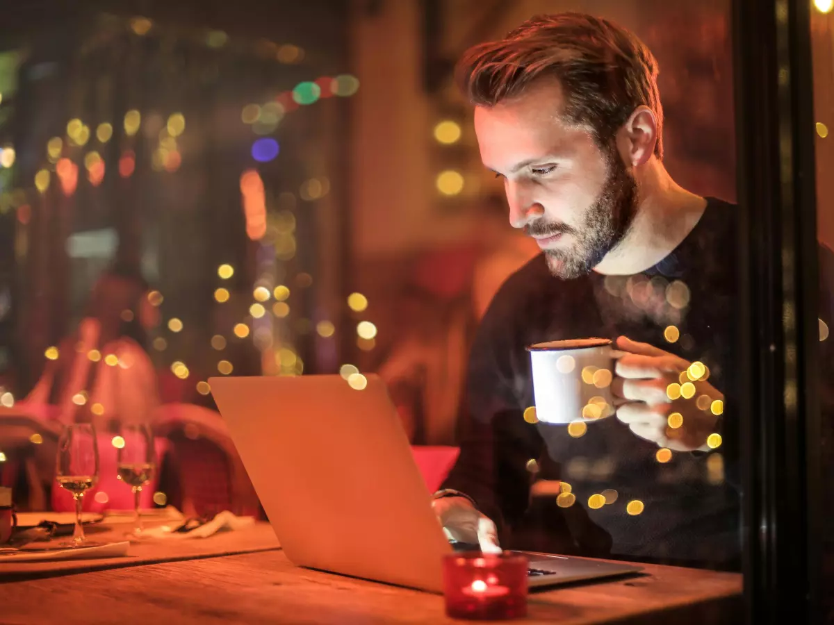 A man with a beard is sitting in a dimly lit cafe, working on his laptop. He has a concerned expression on his face.