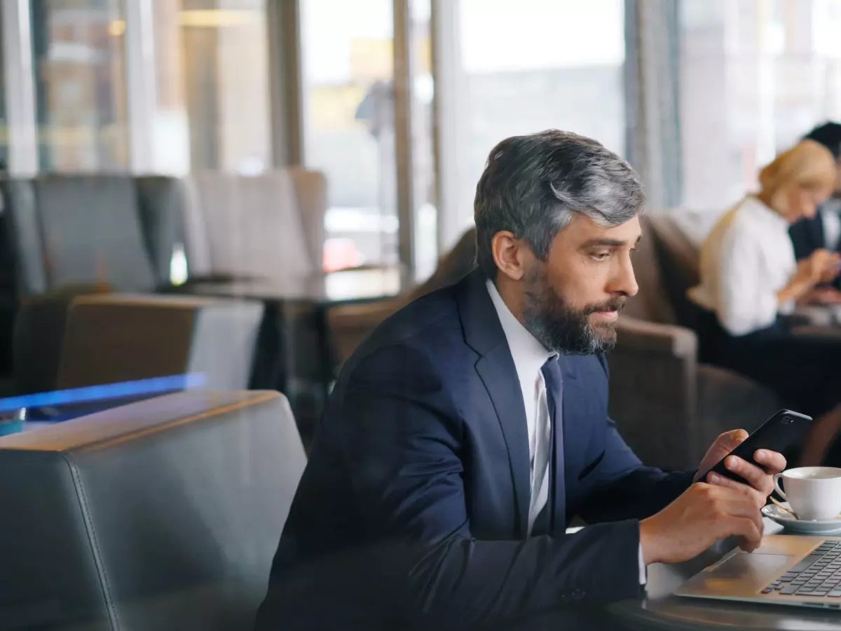 A man in a suit sits at a table in a cafe, looking intently at his phone while a laptop is open in front of him. He is surrounded by other people, suggesting a busy environment.