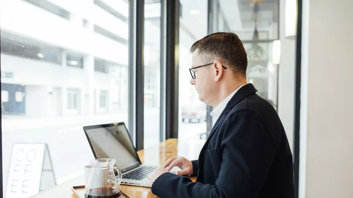 A man in a suit is sitting at a table in front of a laptop. He is looking at the screen and typing on the keyboard. 