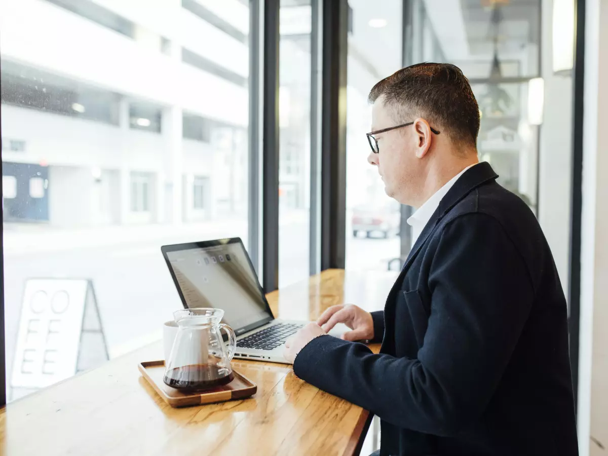 A man in a suit is sitting at a table in front of a laptop. He is looking at the screen and typing on the keyboard. 