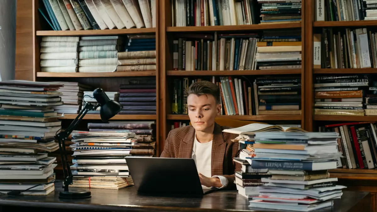 A man in a brown blazer is sitting at a desk in a library, working on a laptop. There are stacks of books and a lamp on the desk. The man is looking down at the laptop, and his expression is focused.