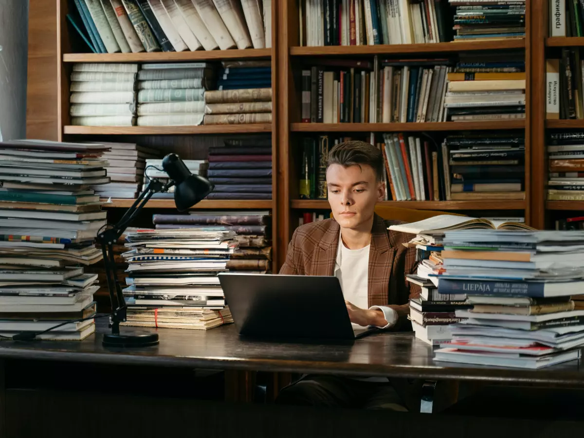 A man in a brown blazer is sitting at a desk in a library, working on a laptop. There are stacks of books and a lamp on the desk. The man is looking down at the laptop, and his expression is focused.