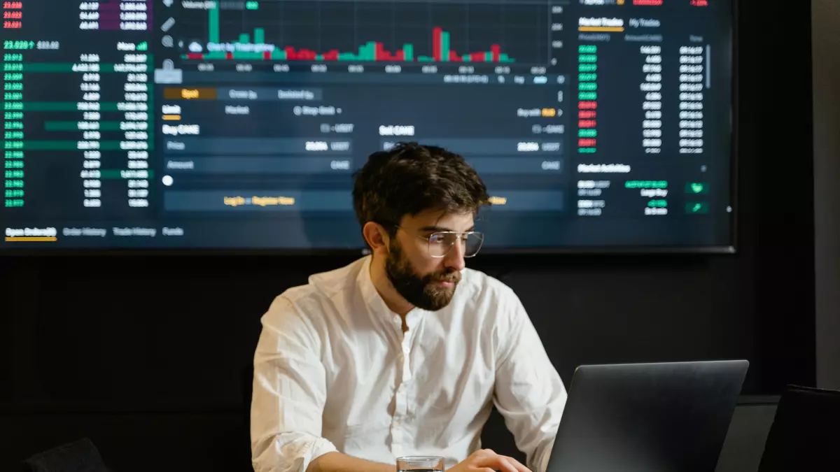 A man sits at a desk in front of a laptop, looking at a large data visualization on a screen behind him.