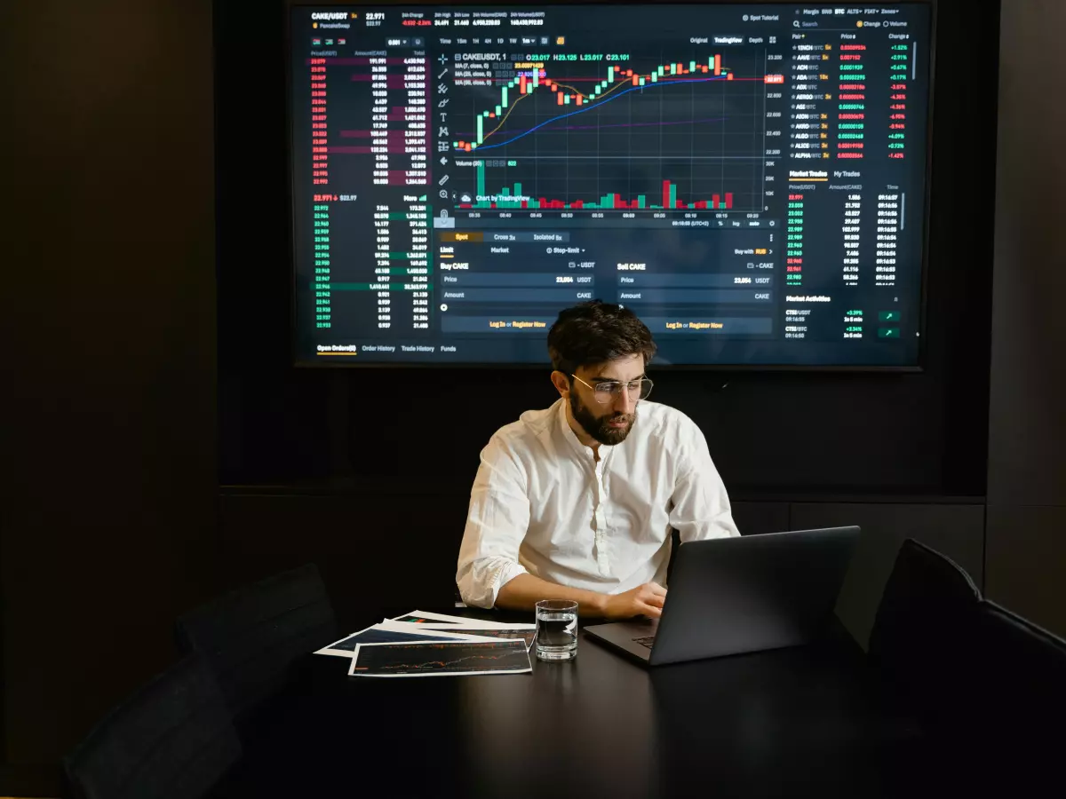 A man sits at a desk in front of a laptop, looking at a large screen with a data chart. He is wearing a white shirt and has a beard.