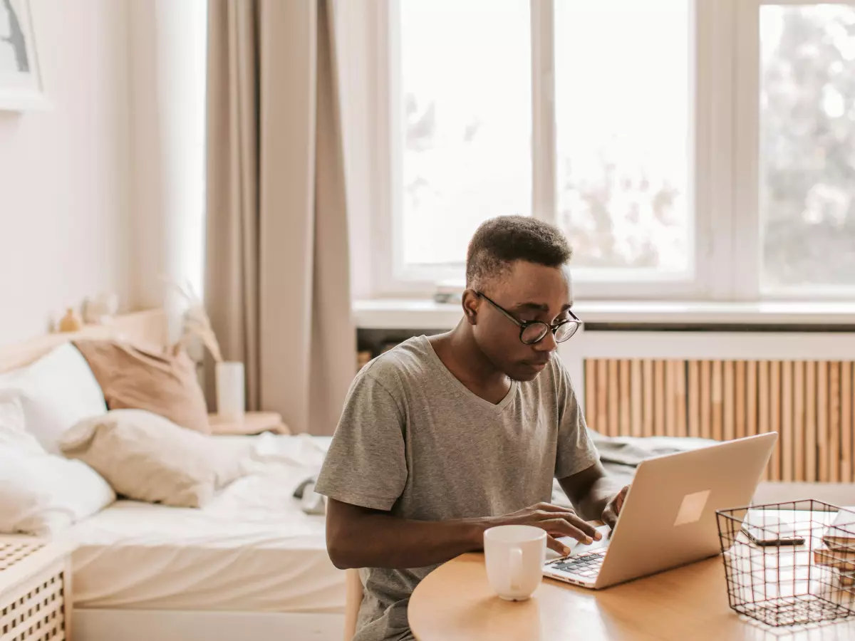 A man sitting at a table, working on his laptop. He is wearing glasses and a grey t-shirt. He is smiling and appears to be focused on his work.